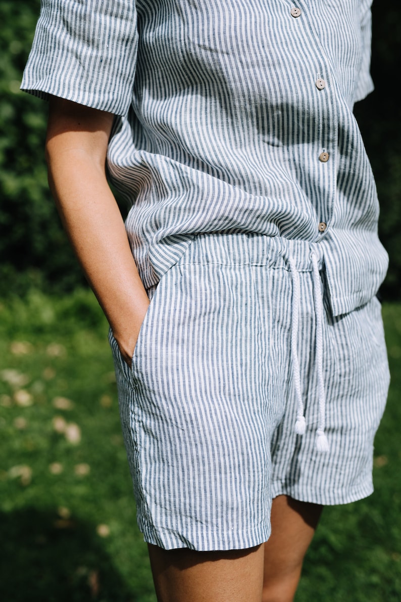 May include: A two-piece linen outfit in a vertical striped pattern of white and green. The outfit includes a short-sleeved button-down shirt and drawstring shorts. The shorts have pockets and a white drawstring. The outfit is made of a lightweight fabric.