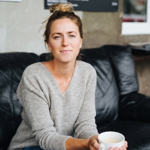 Op de afbeelding: Een vrouw in een grijze trui en blauwe jeans zit op een zwarte leren bank en houdt een witte mok vast.