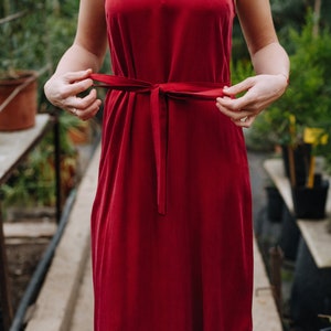 May include: A woman wearing a red sleeveless dress with a self-tie sash at the waist. The dress is a midi length and has a simple, elegant design.