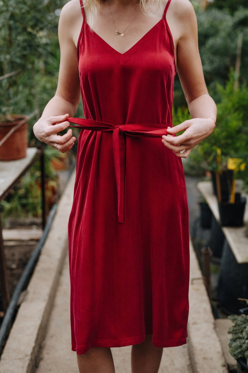 May include: A woman wearing a red sleeveless dress with a self-tie sash at the waist. The dress is a midi length and has a simple, elegant design.