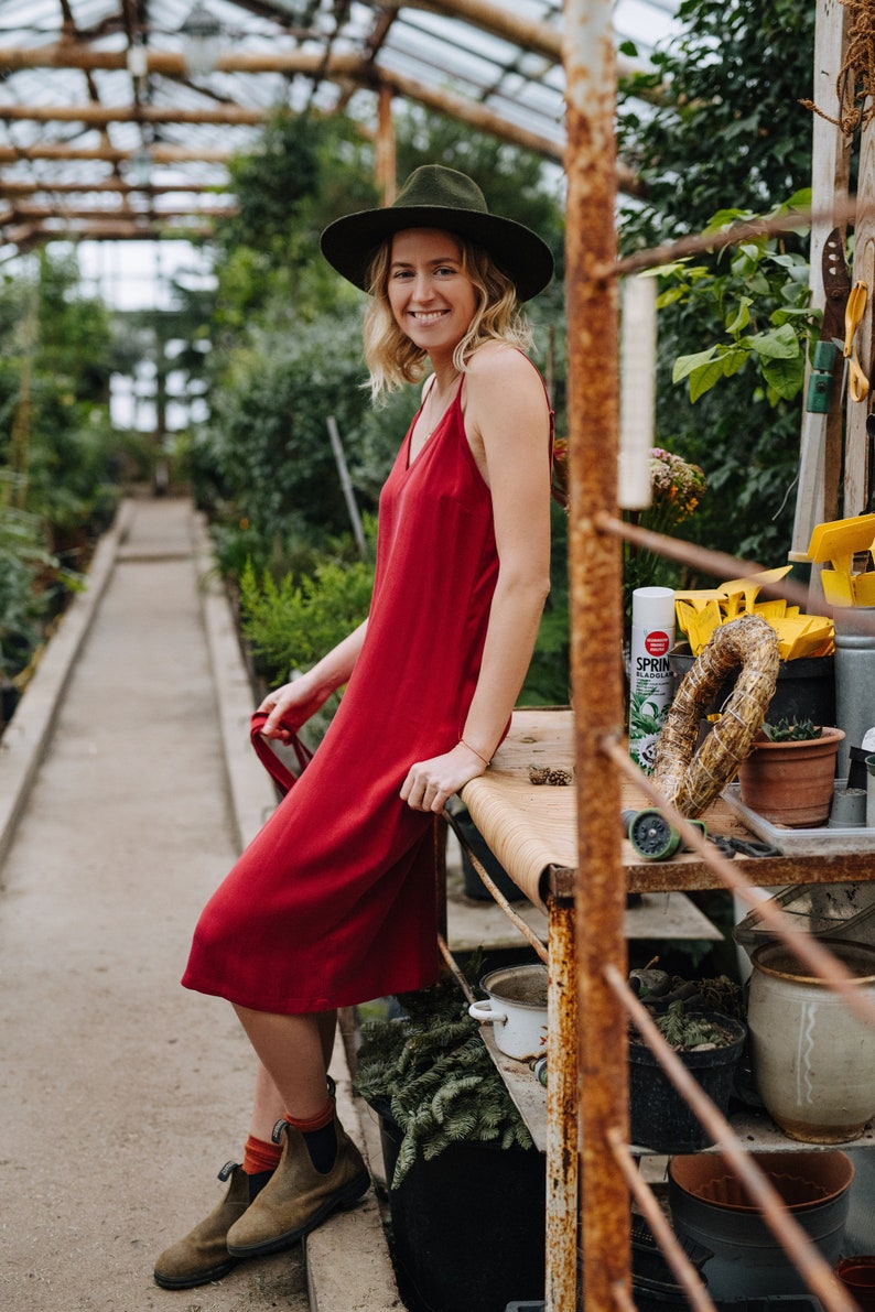 May include: A woman wearing a red dress and a green hat stands in a greenhouse. She is leaning on a rusty metal shelf with plants and gardening supplies. The shelf has a white spray bottle labeled "SPRIN"