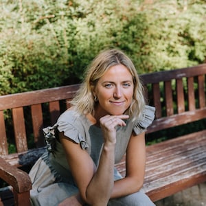 May include: A woman wearing a light gray linen dress with ruffled sleeves sits on a wooden bench outdoors. She is smiling and looking to the side. The background is a blurred green hedge.