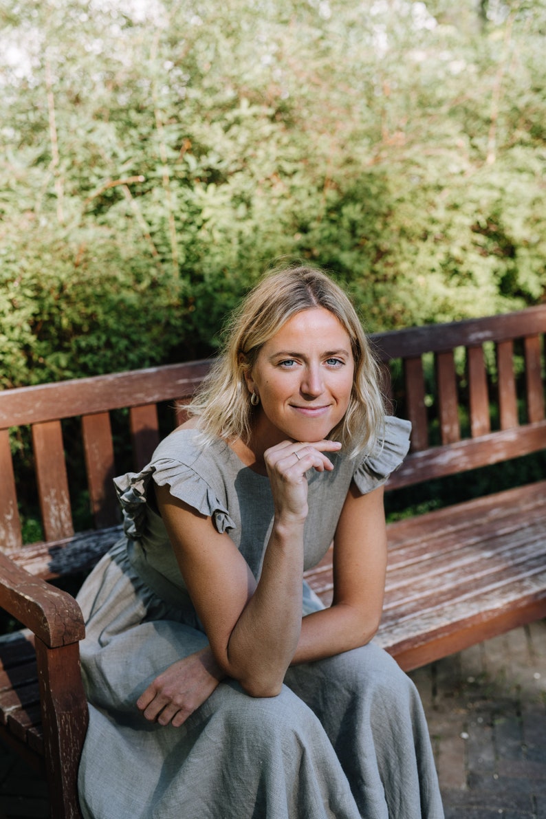 May include: A woman wearing a light gray linen dress with ruffled sleeves sits on a wooden bench outdoors. She is smiling and looking to the side. The background is a blurred green hedge.