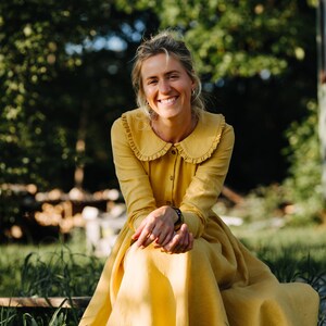 Pode incluir: Uma mulher com um vestido amarelo com colarinho de babados senta-se em um campo gramado. Ela est&aacute; sorrindo e olhando para a c&acirc;mera.