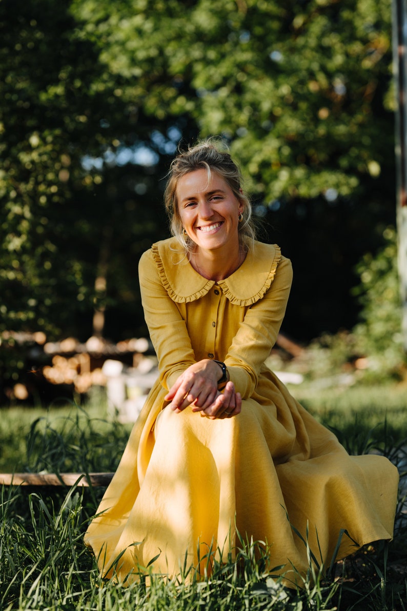 Pode incluir: Uma mulher com um vestido amarelo com colarinho de babados senta-se em um campo gramado. Ela est&aacute; sorrindo e olhando para a c&acirc;mera.