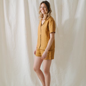 May include: Mustard yellow linen pajama set. The short-sleeved button-down shirt has a black trim and is paired with matching shorts. The outfit is made of a lightweight fabric, perfect for warm weather.