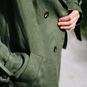 May include: A close-up of a green linen jacket with two buttons and a pocket. The jacket has a textured surface and is made of a natural fabric.