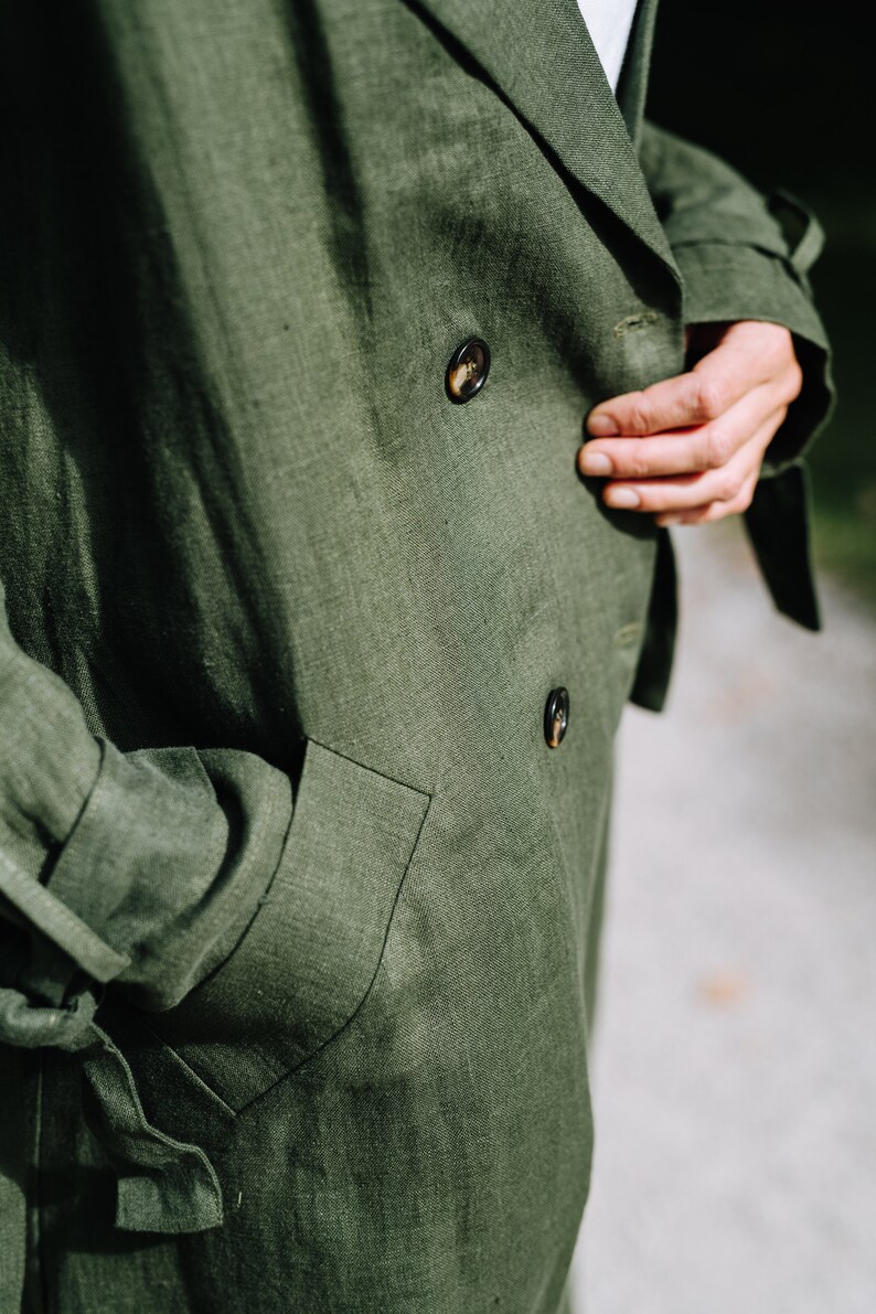 May include: A close-up of a green linen jacket with two buttons and a pocket. The jacket has a textured surface and is made of a natural fabric.