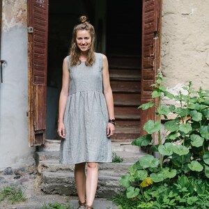 May include: A woman in a gray linen dress stands on a stone path in front of a wooden door. She is smiling and looking at the camera.