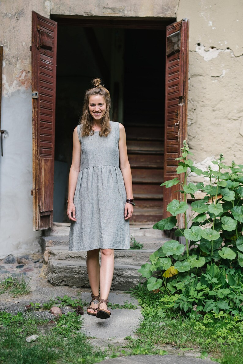 May include: A woman in a gray linen dress stands on a stone path in front of a wooden door. She is smiling and looking at the camera.