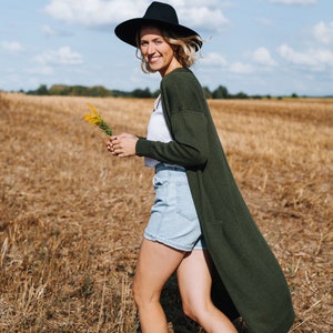 May include: A woman wearing a black hat, a white shirt, a green cardigan, denim shorts, and brown boots walks through a field of dry grass.