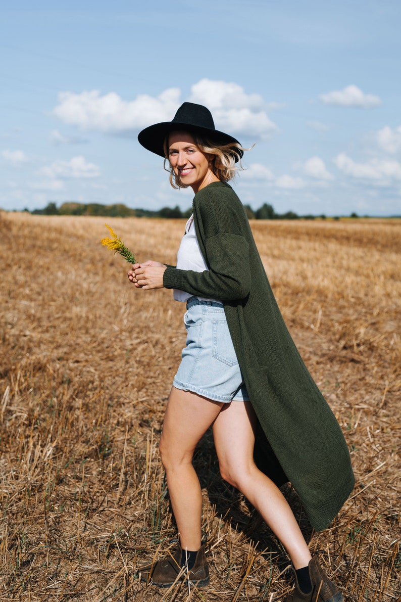 May include: A woman wearing a black hat, a white shirt, a green cardigan, denim shorts, and brown boots walks through a field of dry grass.