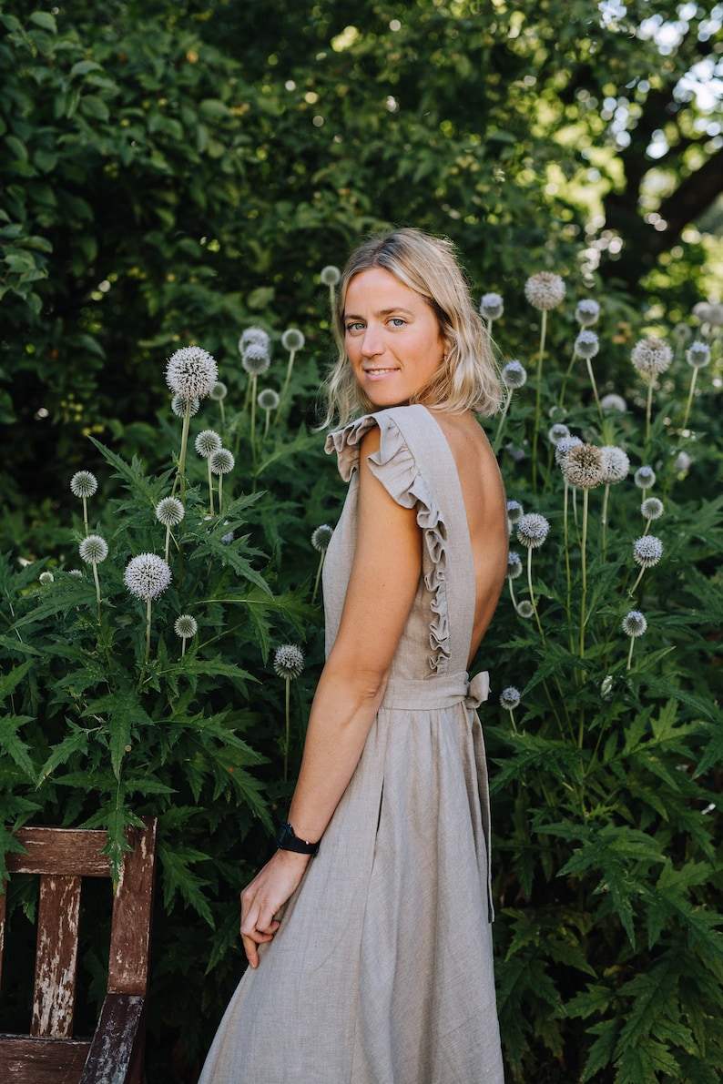 May include: A woman wearing a light brown linen dress with a ruffled neckline and back. The dress has a tie at the back. She is standing in a garden with green foliage and white flowers.