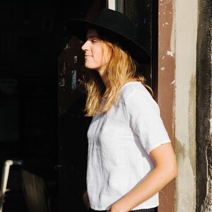 Op de afbeelding: Een vrouw draagt een zwarte fedora, een wit linnen shirt en een zwarte jeans. Ze staat voor een deur met een bruine houten lijst.