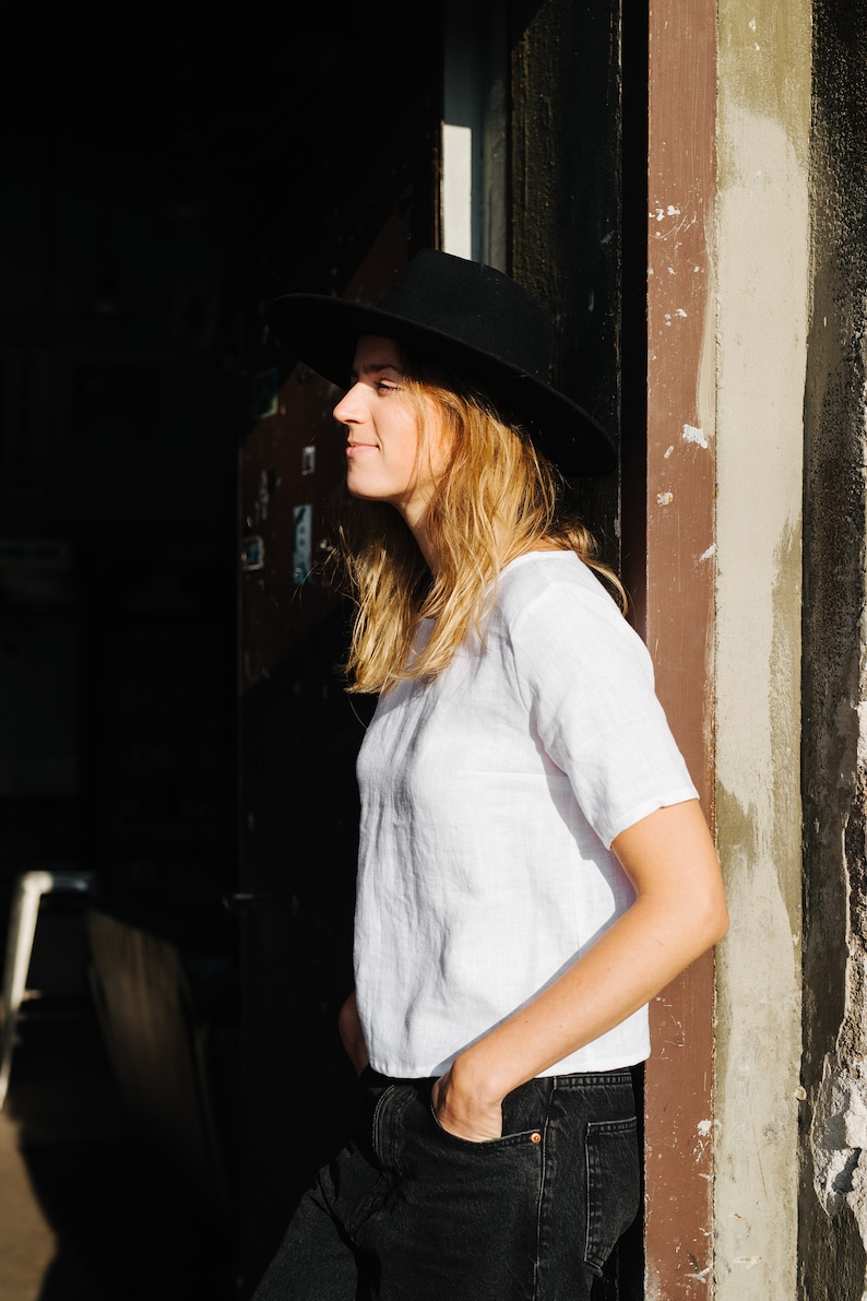Op de afbeelding: Een vrouw draagt een zwarte fedora, een wit linnen shirt en een zwarte jeans. Ze staat voor een deur met een bruine houten lijst.