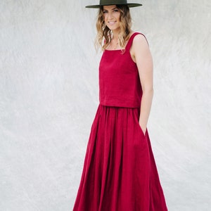 May include: A woman wearing a red linen dress with a wide skirt and a green hat. The dress has a square neckline and thin straps. She is standing in front of a gray wall.