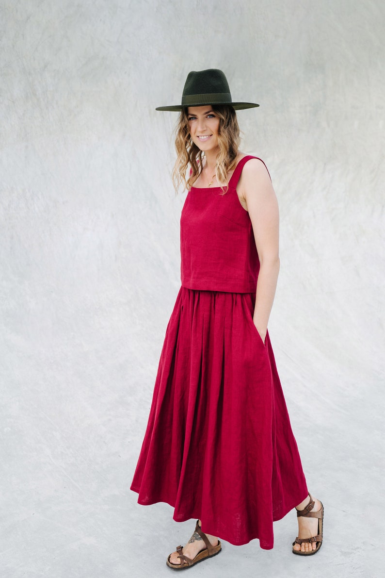 May include: A woman wearing a red linen dress with a wide skirt and a green hat. The dress has a square neckline and thin straps. She is standing in front of a gray wall.
