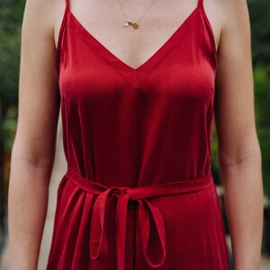 May include: A woman wearing a red satin slip dress with a thin gold chain necklace. The dress has a V-neckline and a tie at the waist.
