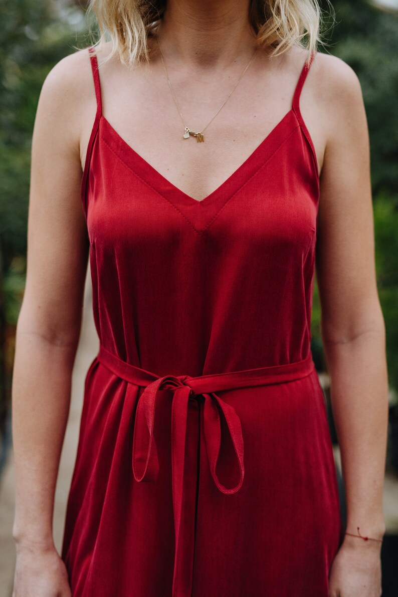 May include: A woman wearing a red satin slip dress with a thin gold chain necklace. The dress has a V-neckline and a tie at the waist.