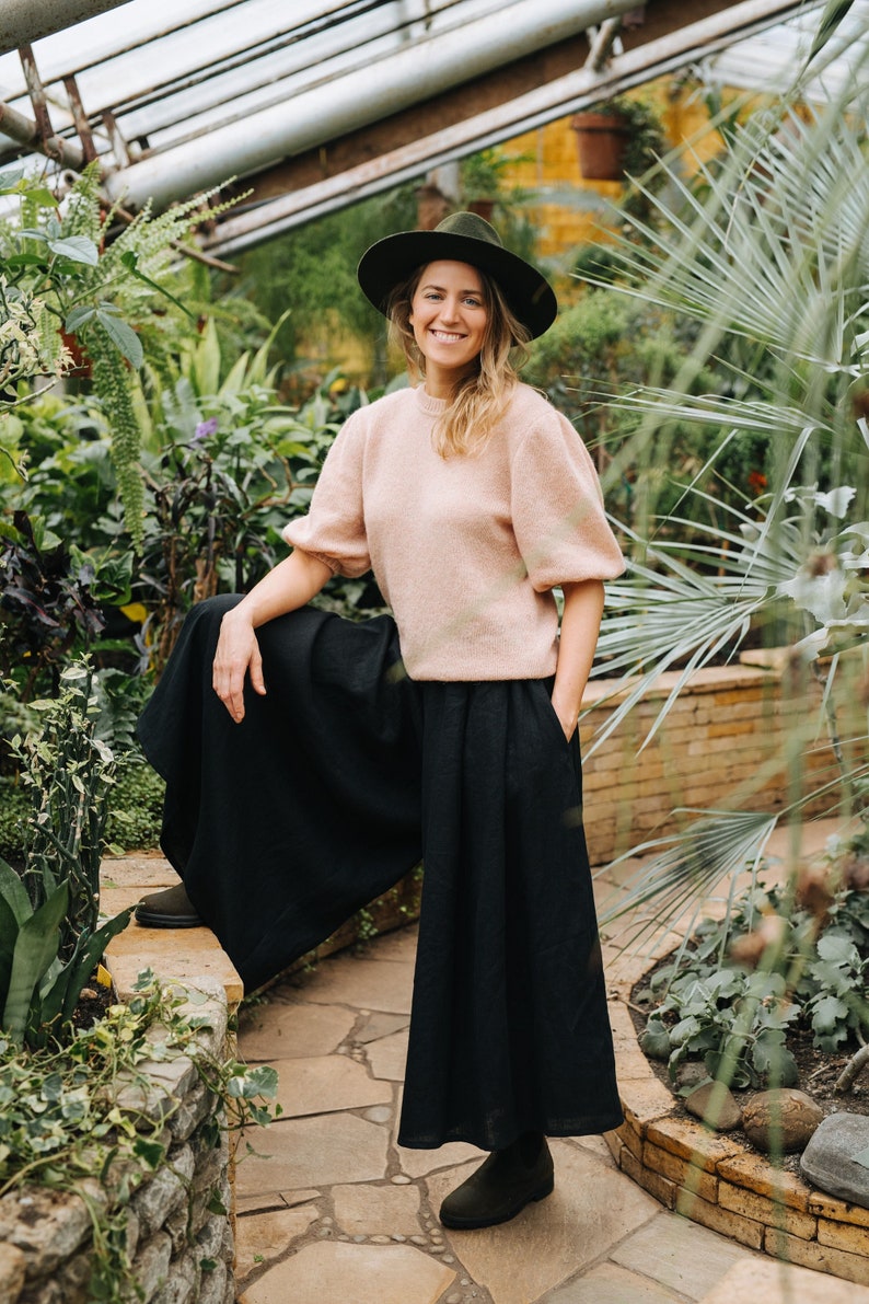 May include: A woman wearing a pink sweater and black wide-leg pants stands in a greenhouse. She is wearing a wide-brimmed hat and smiling at the camera.