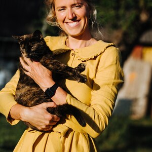 Pode incluir: Uma mulher com um vestido amarelo com colarinho de babados segura um gato de casca de tartaruga em seus bra&ccedil;os. Ela est&aacute; sorrindo para a c&acirc;mera.