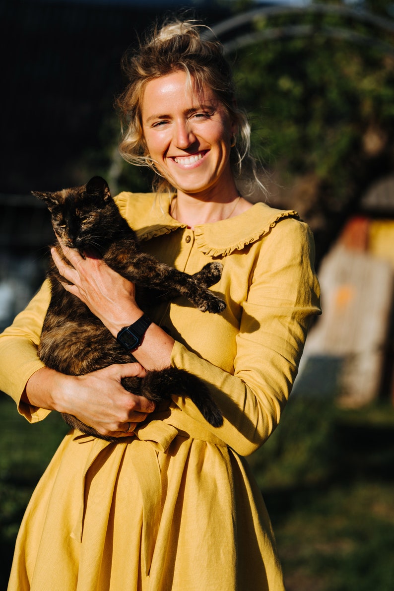 Pode incluir: Uma mulher com um vestido amarelo com colarinho de babados segura um gato de casca de tartaruga em seus bra&ccedil;os. Ela est&aacute; sorrindo para a c&acirc;mera.