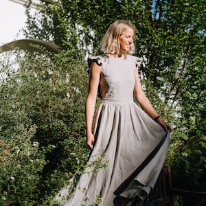 May include: A woman in a light gray linen dress with ruffled straps stands in a garden setting. The dress has a flowy skirt and is sleeveless. She is smiling and looking to the side.