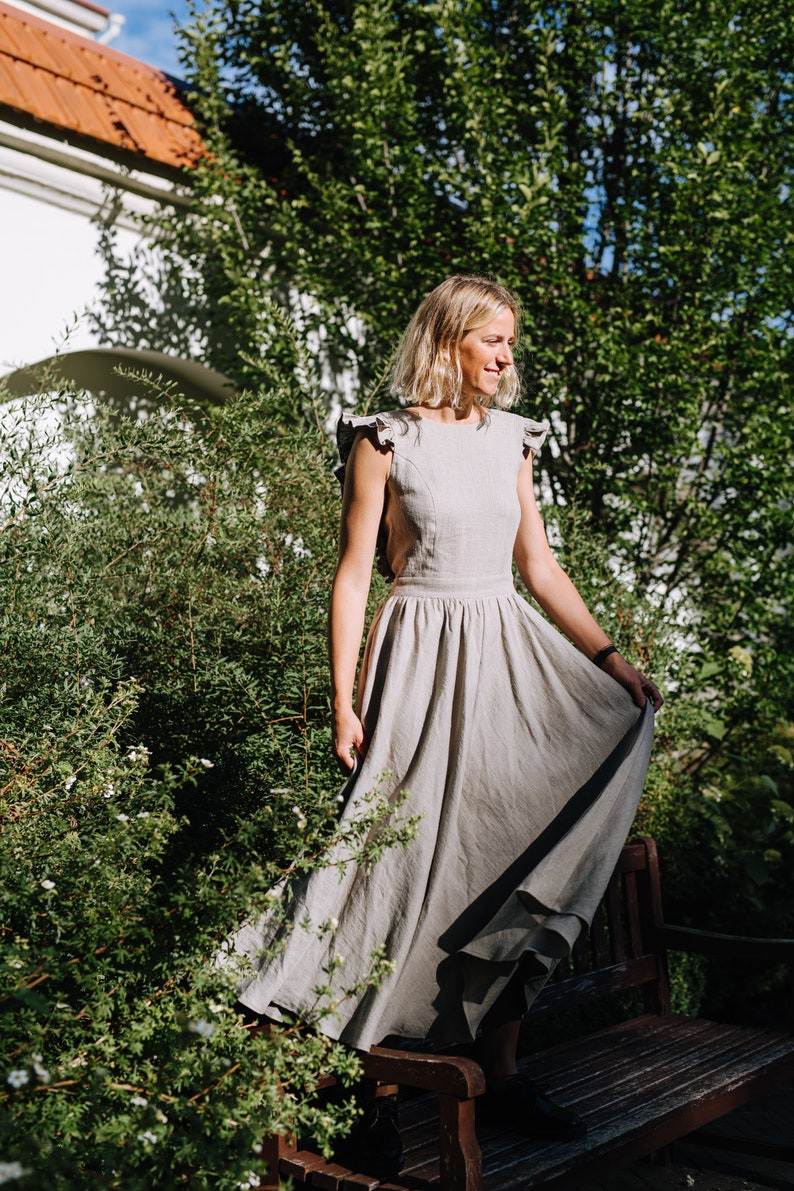 May include: A woman in a light gray linen dress with ruffled straps stands in a garden setting. The dress has a flowy skirt and is sleeveless. She is smiling and looking to the side.