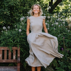 May include: A woman in a light brown linen dress with ruffled sleeves stands in a garden with green foliage and white flowers. She is smiling and her dress is flowing in the wind.