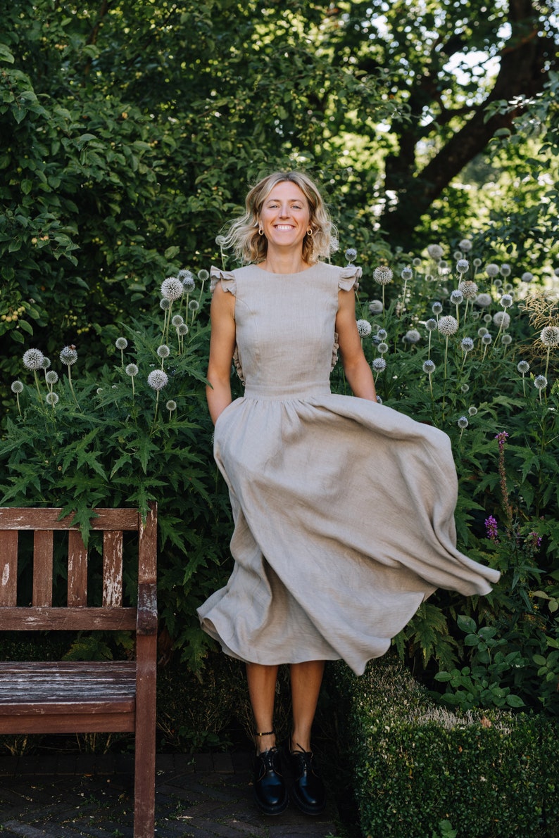 May include: A woman in a light brown linen dress with ruffled sleeves stands in a garden with green foliage and white flowers. She is smiling and her dress is flowing in the wind.
