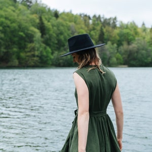 May include: A woman wearing a green linen dress and a black fedora stands by a lake. The dress has a cinched waist and a flowy skirt.
