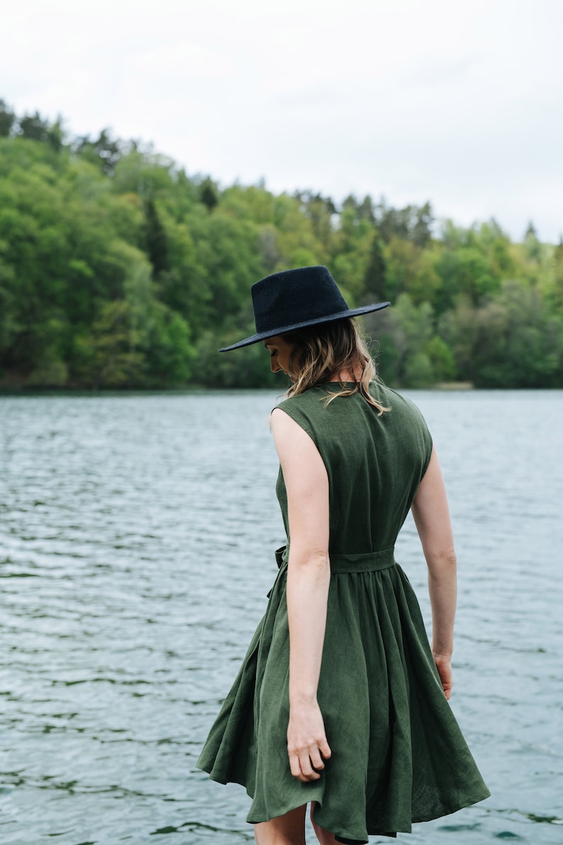 May include: A woman wearing a green linen dress and a black fedora stands by a lake. The dress has a cinched waist and a flowy skirt.