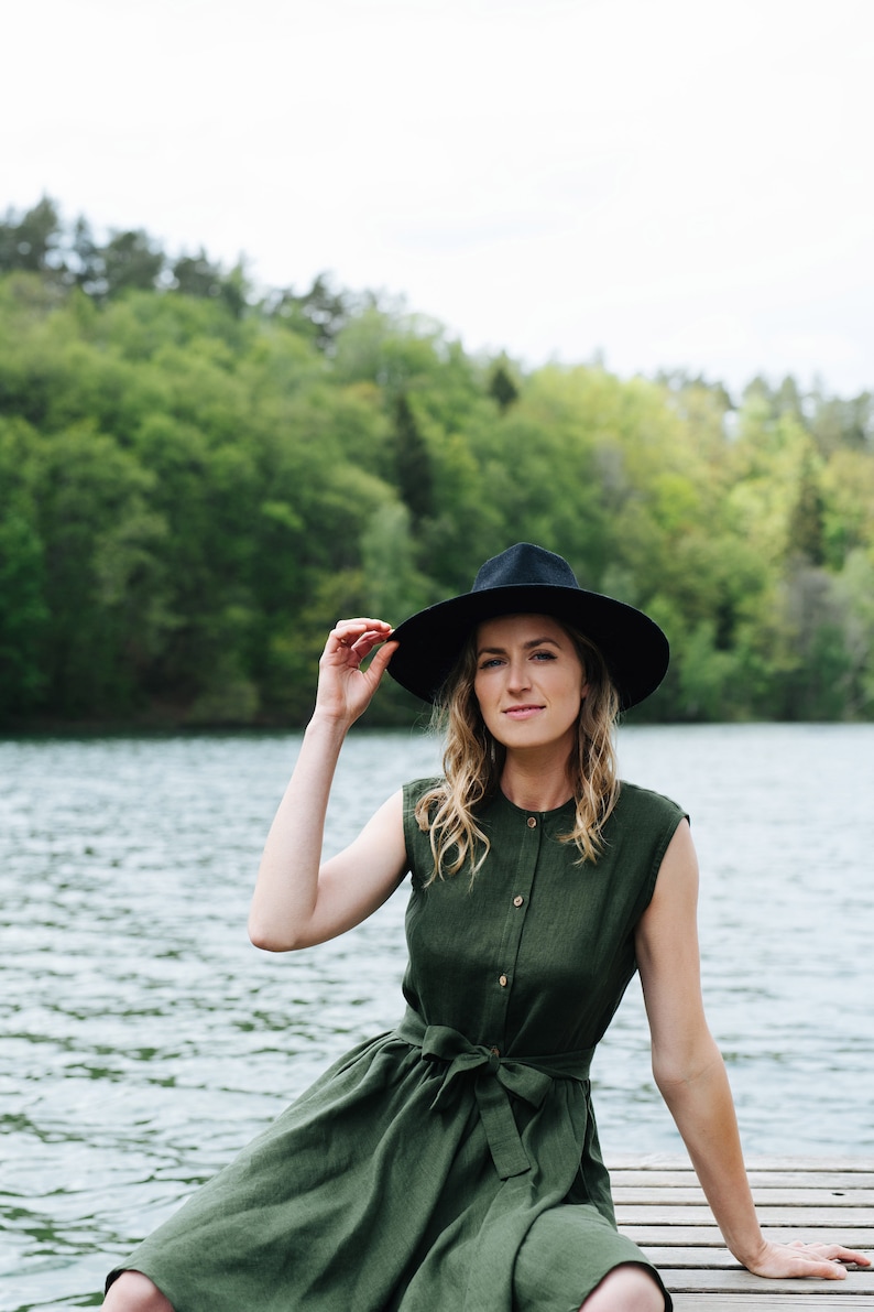May include: A woman wearing a green linen dress with a black hat sits on a wooden dock overlooking a lake. The lake is surrounded by green trees.