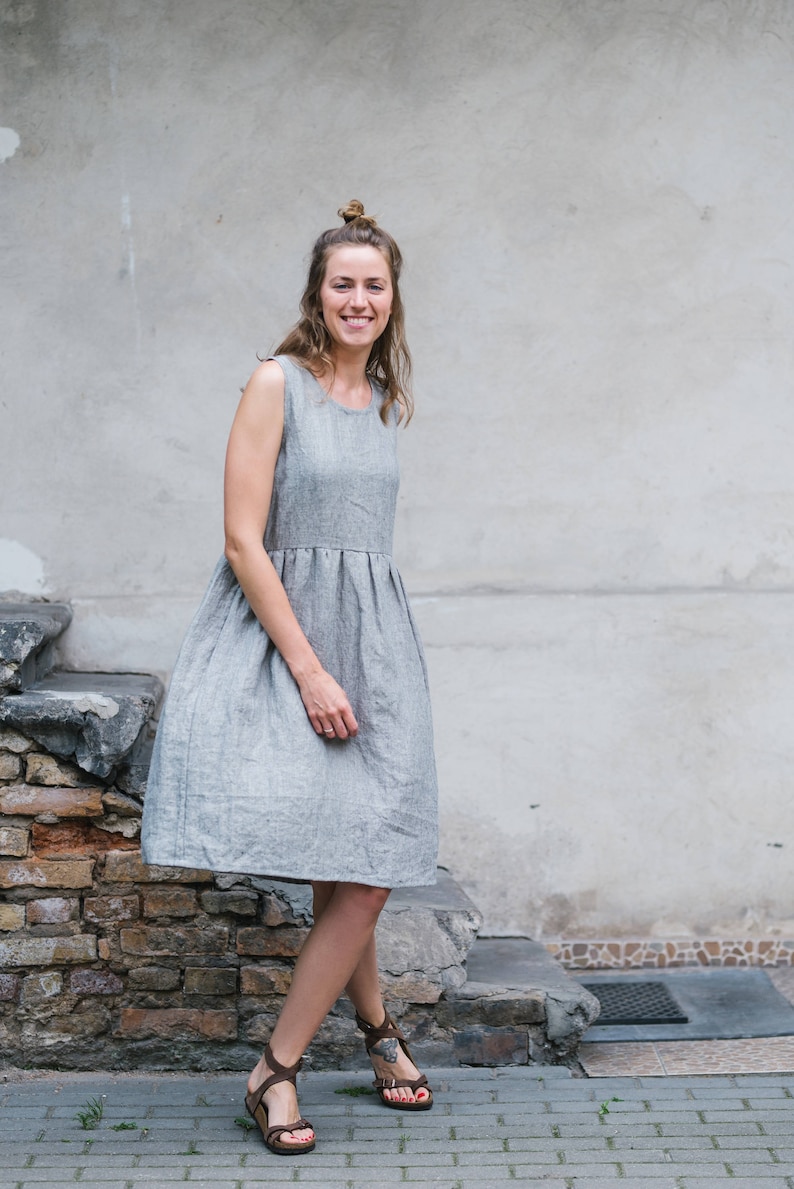 May include: A woman wearing a gray linen dress and brown sandals stands on a brick walkway in front of a gray wall.