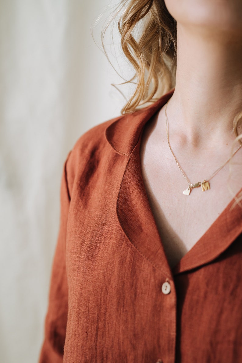 Puede incluir: Primer plano de una persona que lleva una camisa de lino de color &oacute;xido con cuello abotonado. La persona lleva un collar de oro con un dije de coraz&oacute;n y una letra.
