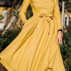 Pode incluir: Um vestido de linho amarelo com colarinho de babados, mangas compridas e cinto de la&ccedil;o. O vestido tem uma saia fluida e &eacute; perfeito para um dia de ver&atilde;o.