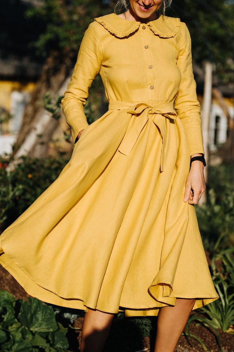 Pode incluir: Um vestido de linho amarelo com colarinho de babados, mangas compridas e cinto de la&ccedil;o. O vestido tem uma saia fluida e &eacute; perfeito para um dia de ver&atilde;o.