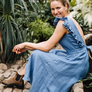 Puede incluir: Una mujer con un vestido de lino azul claro con volantes se sienta en un camino de piedra en un jard&iacute;n. Ella sonr&iacute;e y mira a la c&aacute;mara.