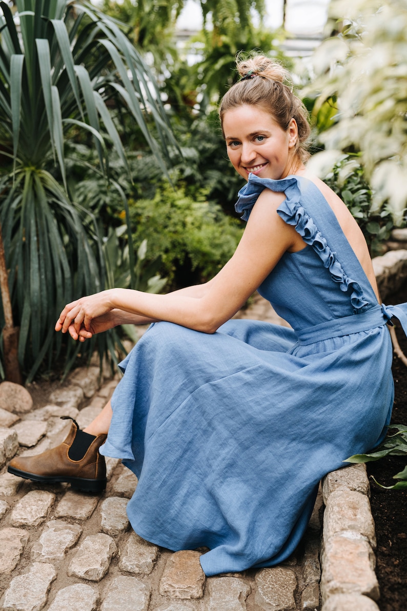 Puede incluir: Una mujer con un vestido de lino azul claro con volantes se sienta en un camino de piedra en un jard&iacute;n. Ella sonr&iacute;e y mira a la c&aacute;mara.