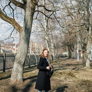 May include: A woman in a black coat and blue jeans stands in a park with bare trees. She is holding a black strap in her hand and looking at the camera.