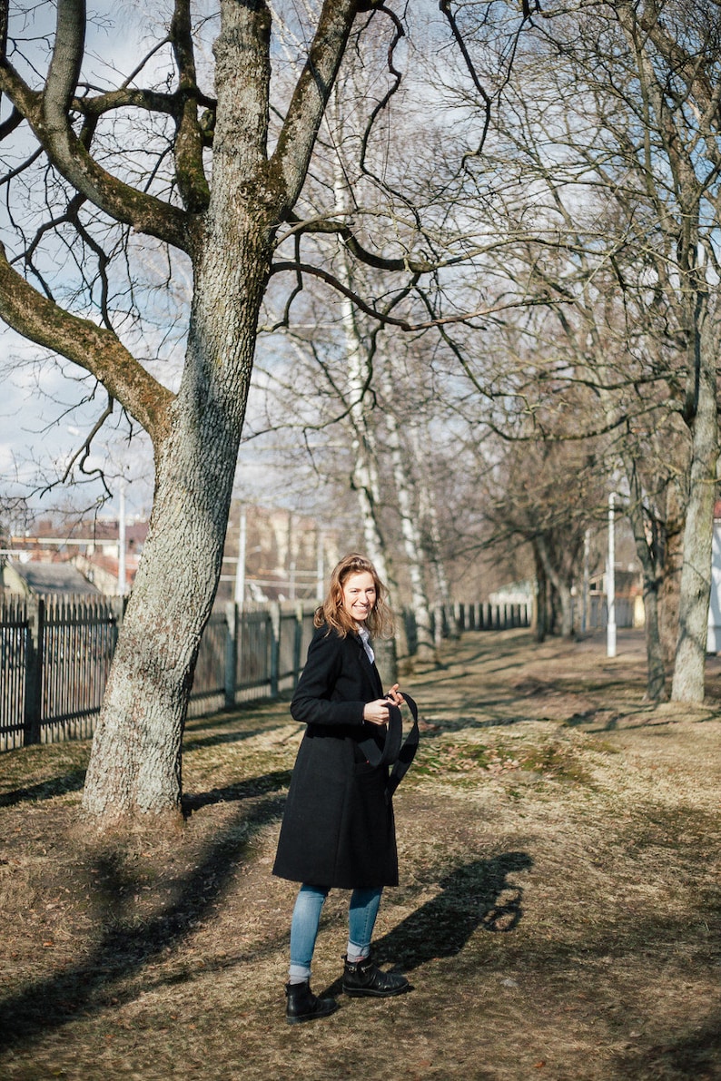 May include: A woman in a black coat and blue jeans stands in a park with bare trees. She is holding a black strap in her hand and looking at the camera.