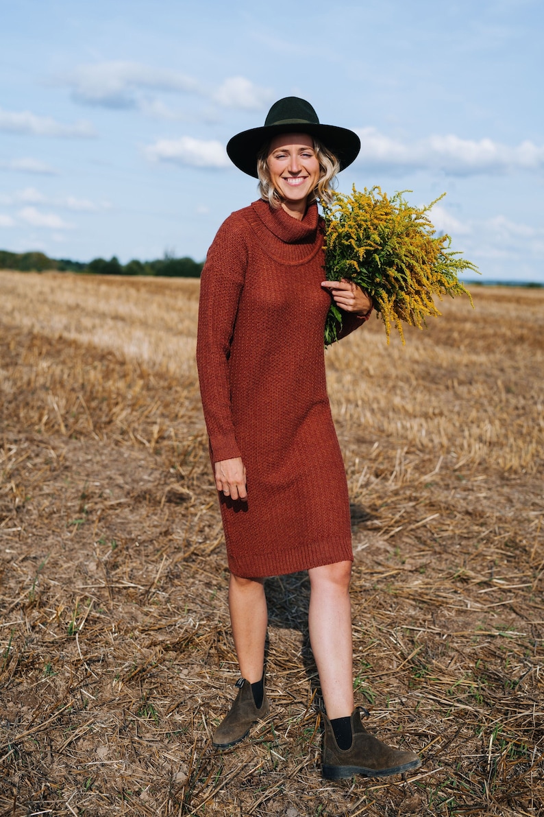 May include: A woman wearing a brown turtleneck sweater dress and a black hat is standing in a field of dry grass. She is holding a bouquet of yellow wildflowers.