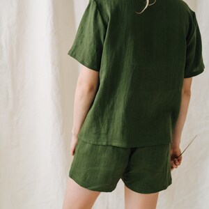 May include: A two-piece olive green linen pajama set. The set includes a short-sleeved button-down shirt and matching shorts. The fabric has a textured appearance. The model is facing away from the camera.