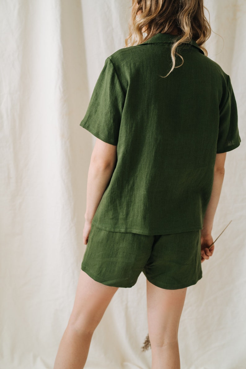May include: A two-piece olive green linen pajama set. The set includes a short-sleeved button-down shirt and matching shorts. The fabric has a textured appearance. The model is facing away from the camera.