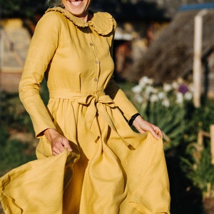 Pode incluir: Uma mulher com um vestido de linho amarelo com colarinho de babados e cinto de la&ccedil;o. Ela est&aacute; sorrindo e girando o vestido.