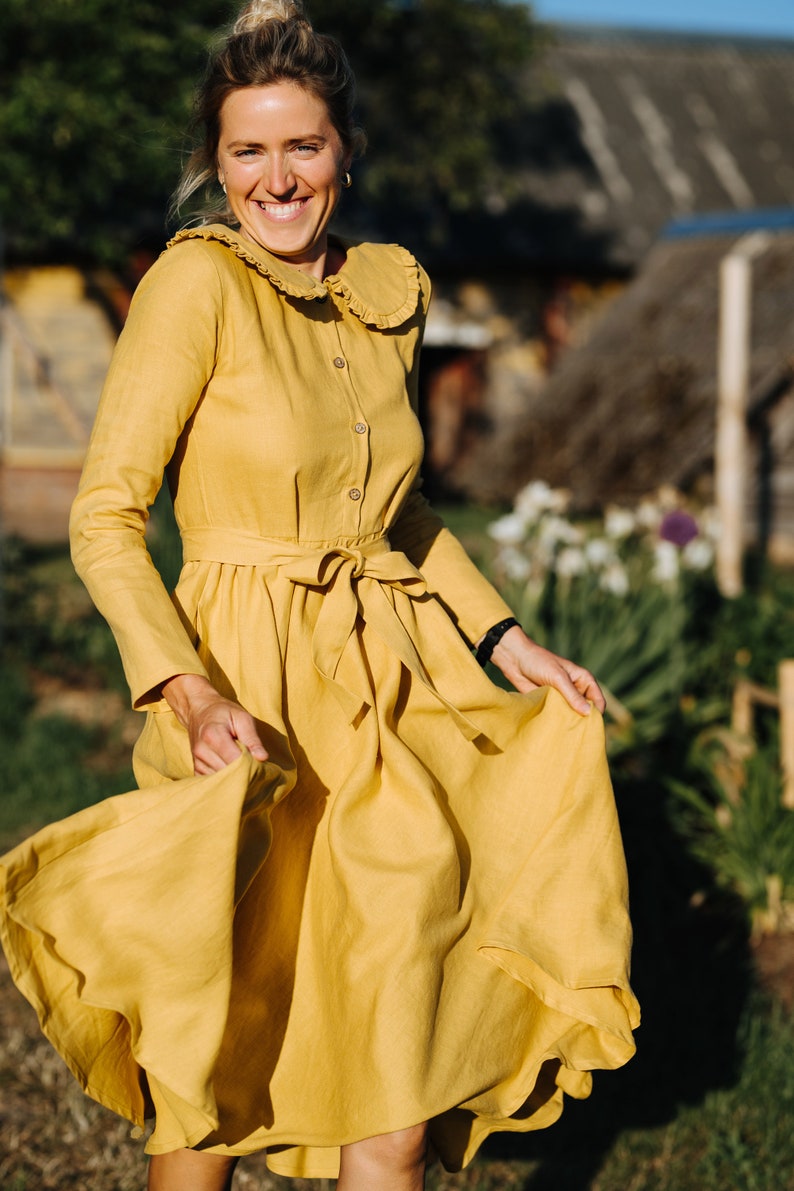 Pode incluir: Uma mulher com um vestido de linho amarelo com colarinho de babados e cinto de la&ccedil;o. Ela est&aacute; sorrindo e girando o vestido.