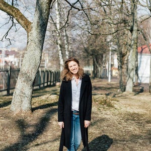 May include: A woman wearing a black coat, blue jeans, and black boots is standing in a park. She is smiling and looking at the camera.