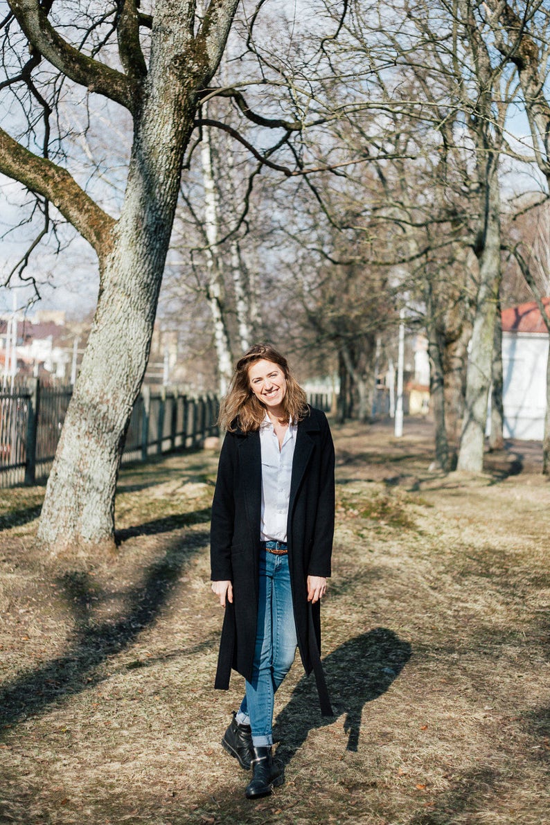 May include: A woman wearing a black coat, blue jeans, and black boots is standing in a park. She is smiling and looking at the camera.