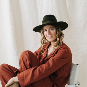 Puede incluir: Una mujer con un mono de lino color &oacute;xido y un sombrero fedora verde oscuro se sienta en una silla blanca.
