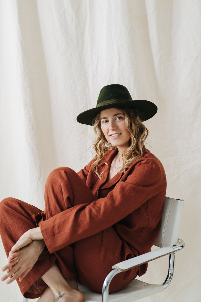 Puede incluir: Una mujer con un mono de lino color &oacute;xido y un sombrero fedora verde oscuro se sienta en una silla blanca.